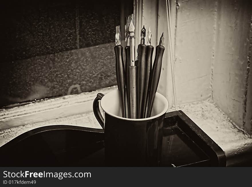 Artists cup full of old dip pens sits on wooden desk in this toned image. Artists cup full of old dip pens sits on wooden desk in this toned image