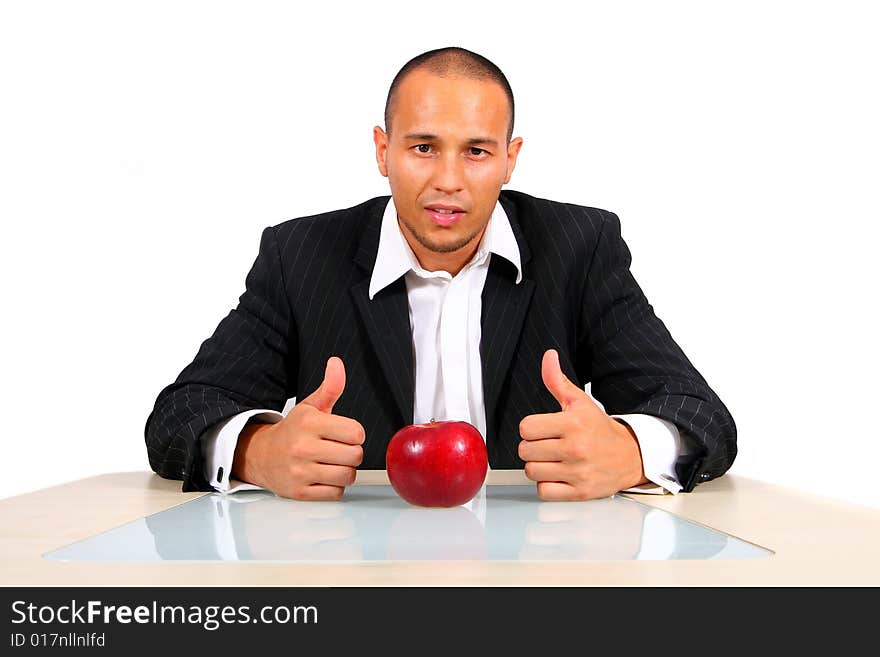 Young businessman sitting in front of a red apple thinking with his thumbs up. Isolate over white. The table mirrors the image creating a nice effect!. Young businessman sitting in front of a red apple thinking with his thumbs up. Isolate over white. The table mirrors the image creating a nice effect!