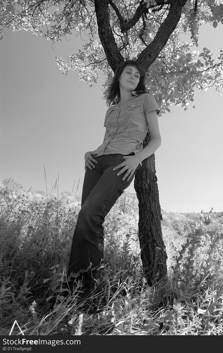 The girl in the field leans against a tree. Monochrome