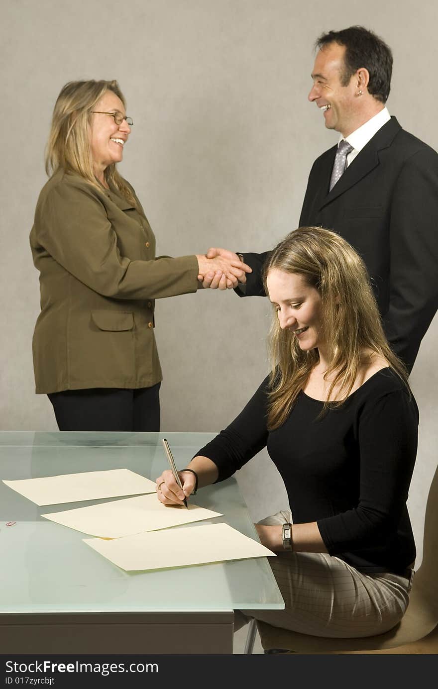 Three people are in a room together. The youngest member is smiling and studying paper on the table in front of her. The other two are shaking hands and smiling at each other. Vertically framed shot. Three people are in a room together. The youngest member is smiling and studying paper on the table in front of her. The other two are shaking hands and smiling at each other. Vertically framed shot.