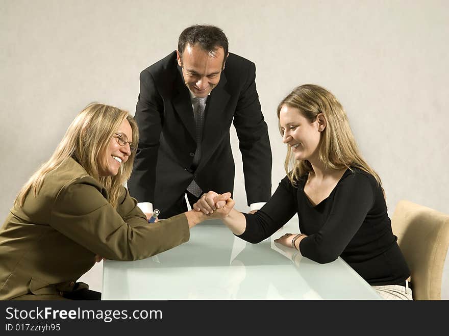 Two women arm wrestling across a table while a man stands in the middle watching. Horizontally framed photo.