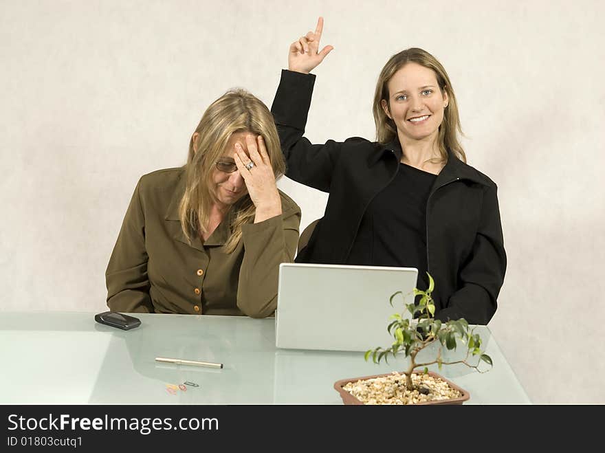 Two Women are sitting at a table in a business meeting.  One woman has her eyes closed and looks stressed.  The other woman is smiling and has her hand pointing upwards.  Horizontally framed shot. Two Women are sitting at a table in a business meeting.  One woman has her eyes closed and looks stressed.  The other woman is smiling and has her hand pointing upwards.  Horizontally framed shot.