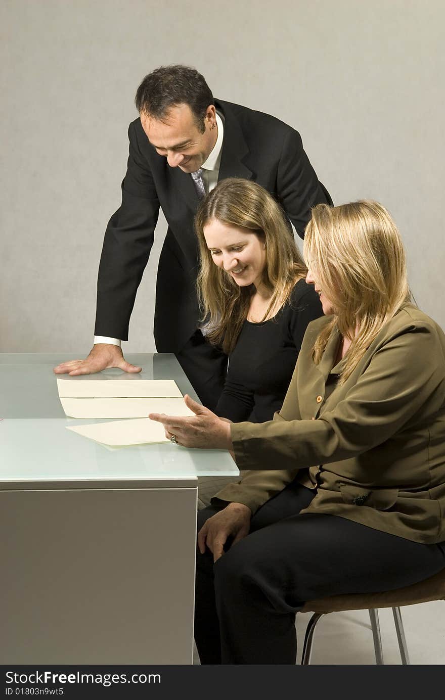 Three people are in a business meeting. They are smiling and looking at some pieces of paper on the table. The women are sitting and the man is standing. Vertically framed shot. Three people are in a business meeting. They are smiling and looking at some pieces of paper on the table. The women are sitting and the man is standing. Vertically framed shot.