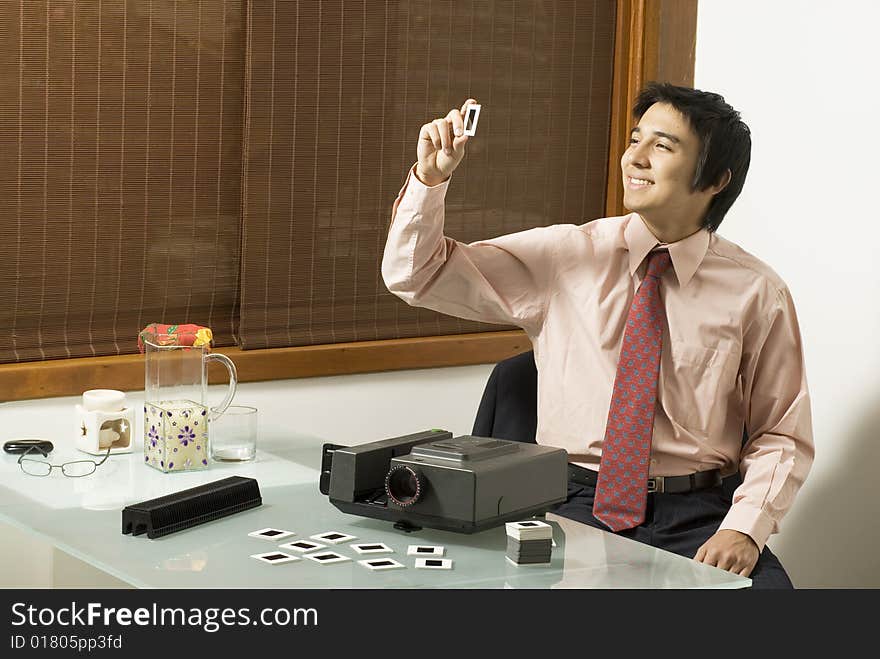 Man smiling as he looks at a slide from a slide show projector. There is a juice pitcher, a candle, and a projector on the table where he is seated. Horizontally framed photo. Man smiling as he looks at a slide from a slide show projector. There is a juice pitcher, a candle, and a projector on the table where he is seated. Horizontally framed photo.