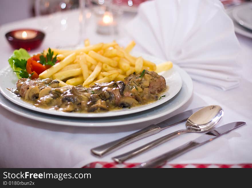 French fries with mushrooms and pork meat in the background are candles, cutlery and napkins. French fries with mushrooms and pork meat in the background are candles, cutlery and napkins