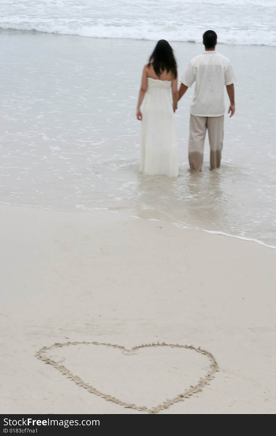 Heart in the sand with bride and groom walking towards the ocean. Heart in the sand with bride and groom walking towards the ocean.