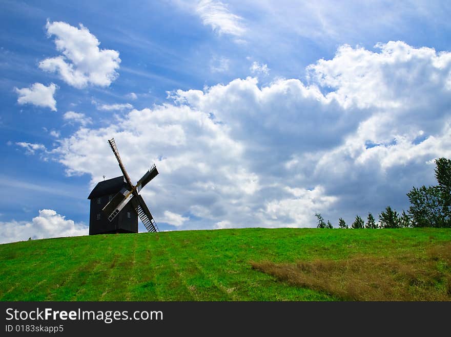 Wooden windmill against the summer blue sky with white clouds on a green hill with a birch