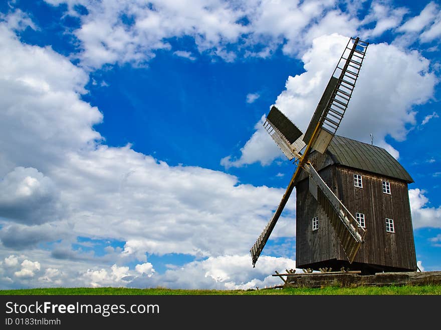 Wooden windmill against the summer blue sky with white clouds on a green hill with a birch