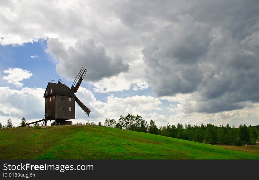 Wooden windmill against the summer blue sky with white clouds on a green hill with a birch