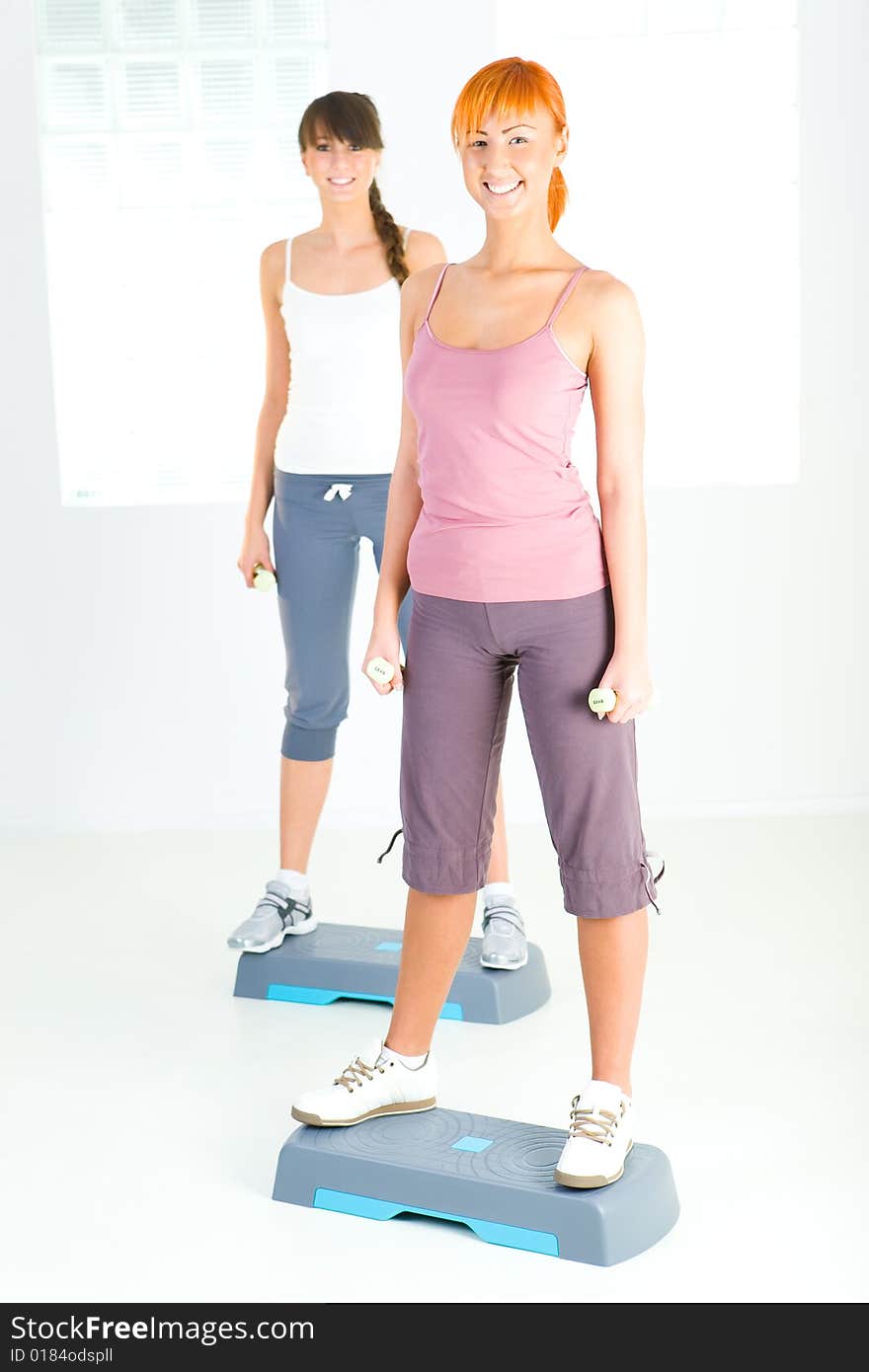 Two young women doing exercise on aerobic step. They're looking at camera. Front view. Two young women doing exercise on aerobic step. They're looking at camera. Front view.