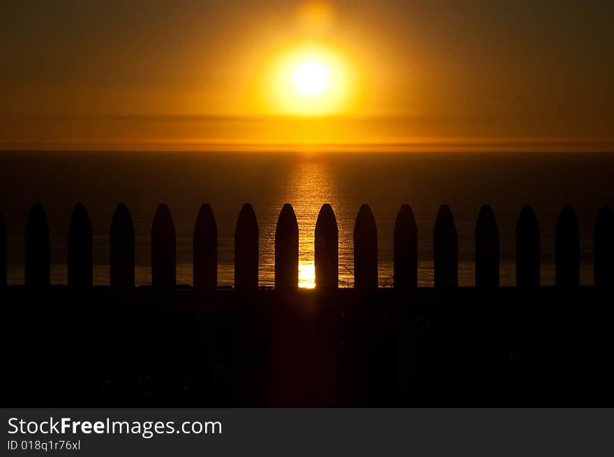 The sun sets on the Pacific Ocean over a white picket fence, silhouetted by the sun. The sun sets on the Pacific Ocean over a white picket fence, silhouetted by the sun.