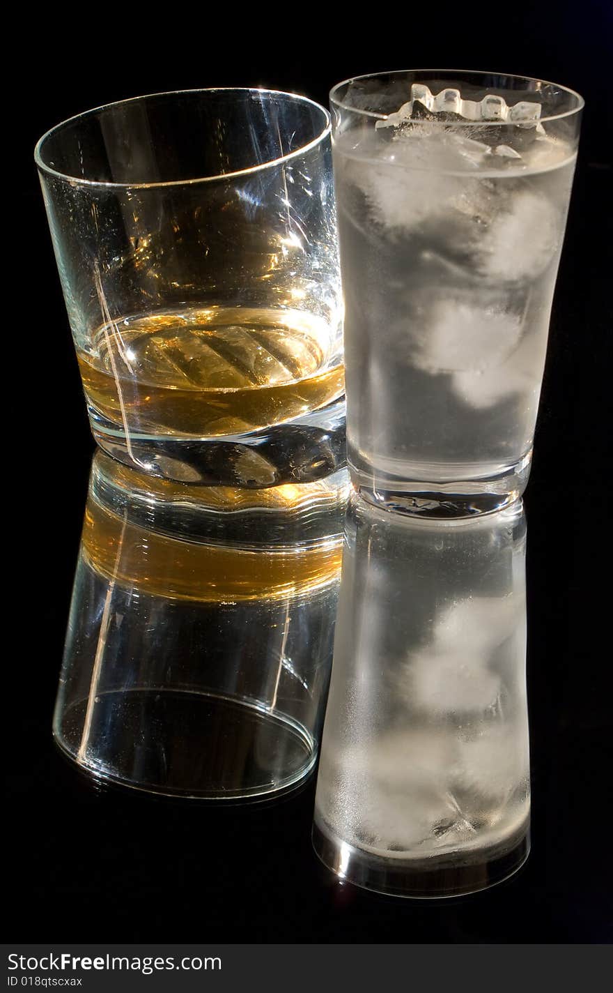 A glass of whiskey and a glass of ice water against a black background, reflected in the surface. A glass of whiskey and a glass of ice water against a black background, reflected in the surface.