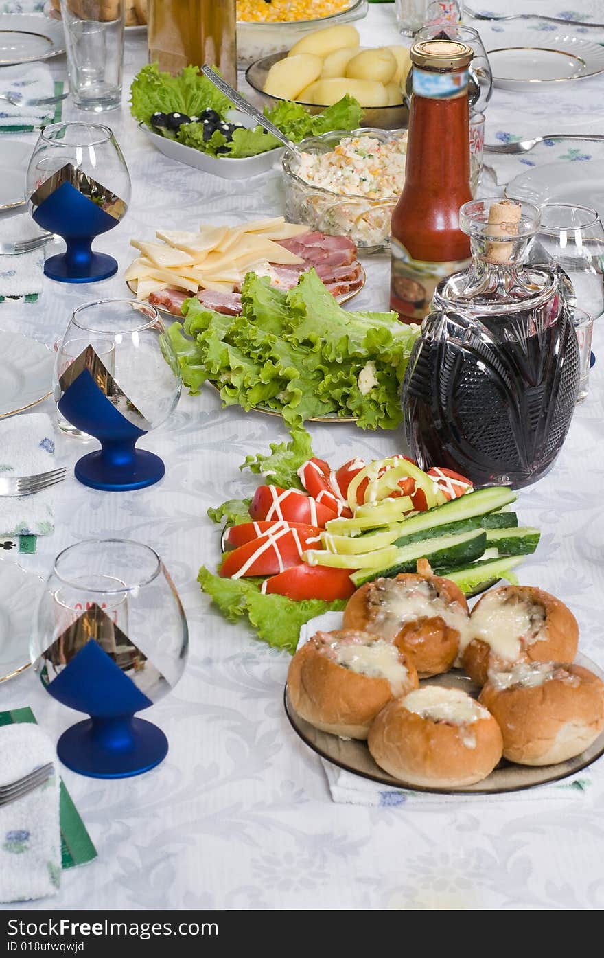 Tablewares and meal on a dining table. Meal, potato, tomatoes, cucumbers, pepper, greens, salad, bread, rolls, meat, cheese, olives, red wine, a tomato sauce, ketchup, tablewares, bottles, plates, plugs, wine glasses, glasses, wine-glasses, a white cloth,. Tablewares and meal on a dining table. Meal, potato, tomatoes, cucumbers, pepper, greens, salad, bread, rolls, meat, cheese, olives, red wine, a tomato sauce, ketchup, tablewares, bottles, plates, plugs, wine glasses, glasses, wine-glasses, a white cloth,