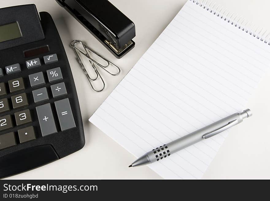 A desktop calculator, stapler and paper clips stands next to a blank writing pad with a stylish pen atop. A desktop calculator, stapler and paper clips stands next to a blank writing pad with a stylish pen atop