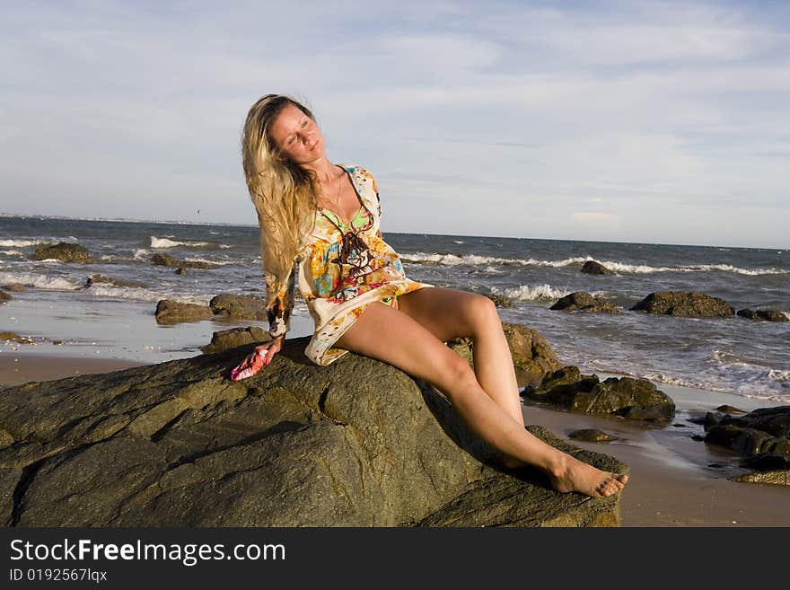 Young blonde woman sitting on a big stone near an ocean at sunset time.