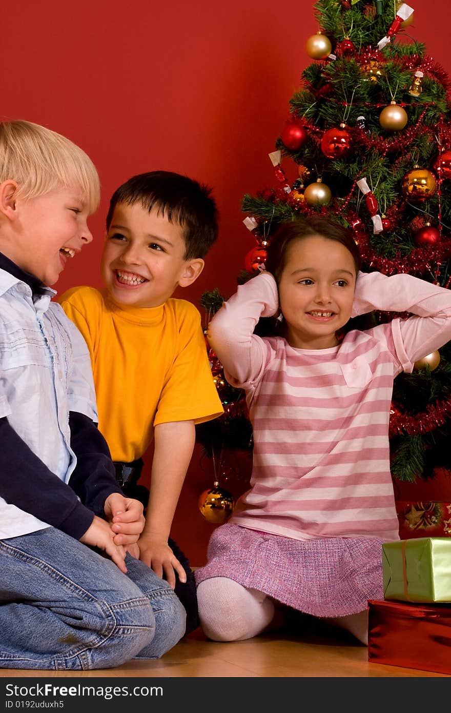 Smiling children sitting by Christmas tree in red room