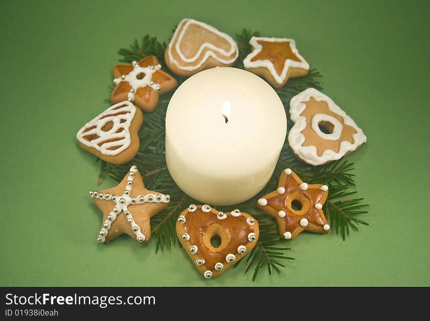 White pillar candle on fir branches surrounded by decorated gingerbread cookies on green background. White pillar candle on fir branches surrounded by decorated gingerbread cookies on green background