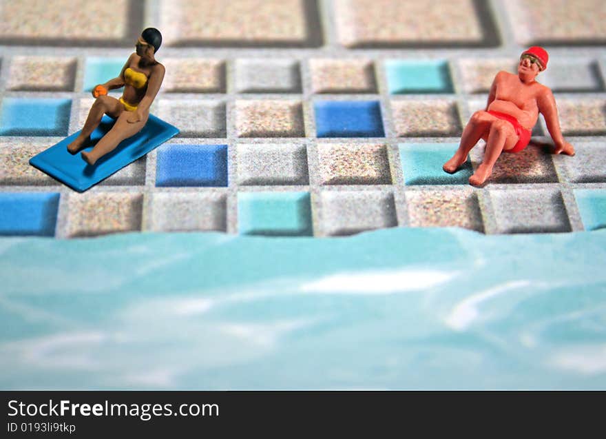 A darker-skinned woman is sunbathing near a sunburned overweight man. They are on a tile patio by a pool. They are miniature figures. Copyspace at bottom. A darker-skinned woman is sunbathing near a sunburned overweight man. They are on a tile patio by a pool. They are miniature figures. Copyspace at bottom.