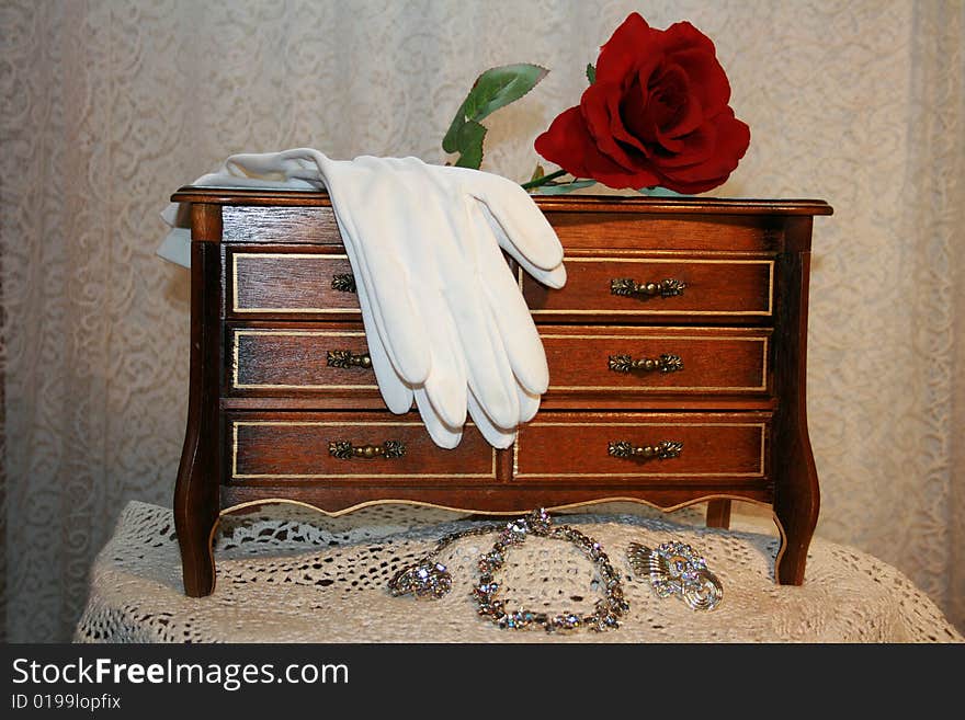 A jewelry box with white gloves and a red rose on it and a necklace and earrings in front of it in front of a lace background.