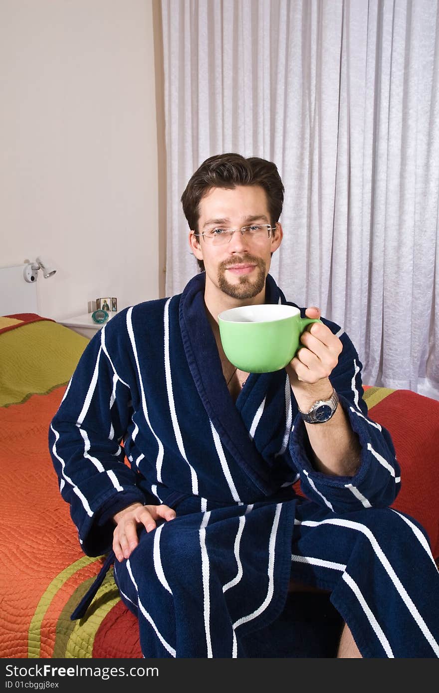 Young man in glasses wearing blue striped bathrobe sitting in his bedroom and drinking coffee or tea from green cup. Young man in glasses wearing blue striped bathrobe sitting in his bedroom and drinking coffee or tea from green cup