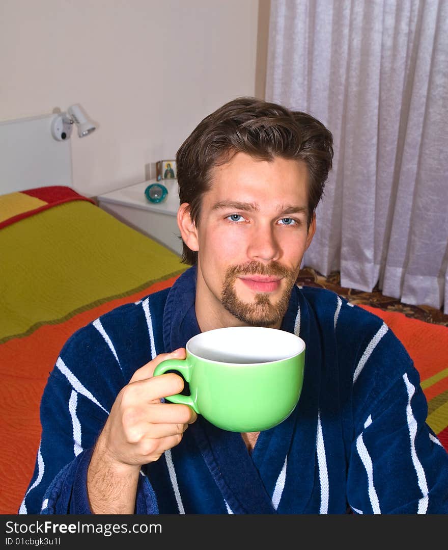 Young man in glasses wearing blue striped bathrobe sitting in his bedroom and drinking coffee or tea from green cup. Young man in glasses wearing blue striped bathrobe sitting in his bedroom and drinking coffee or tea from green cup