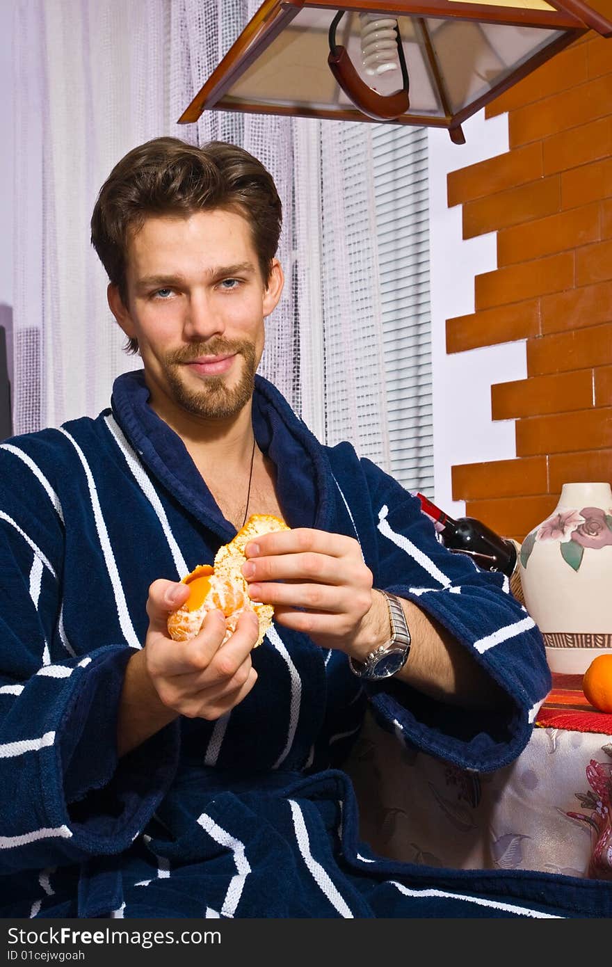 Young man in bathrobe in kitchen peels tangerine