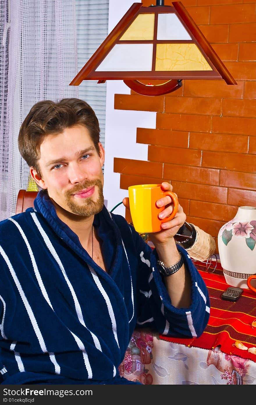 Young man wearing blue striped bathrobe sitting in his kitchen and drinking his breakfast tea. Young man wearing blue striped bathrobe sitting in his kitchen and drinking his breakfast tea