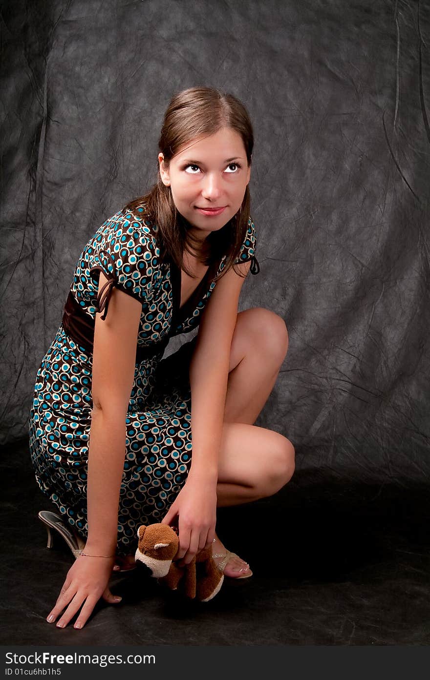 Dark haired girl sitting on hunkers with hands put down and looking up with teddy bear on hand and smiling on black background