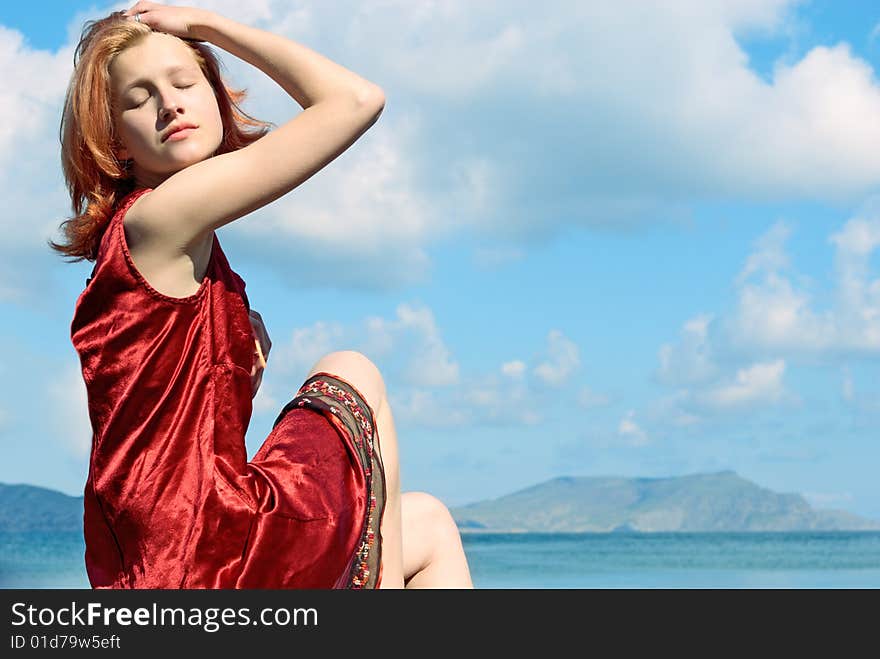 Girl in red dress sitting beside the sea on the blue sky background.