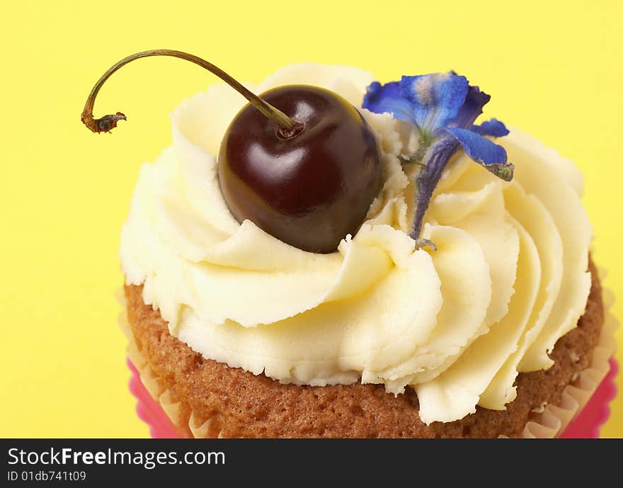 Miniature vanilla cupcake with icing, fresh cherry and blue flower on yellow background. Miniature vanilla cupcake with icing, fresh cherry and blue flower on yellow background