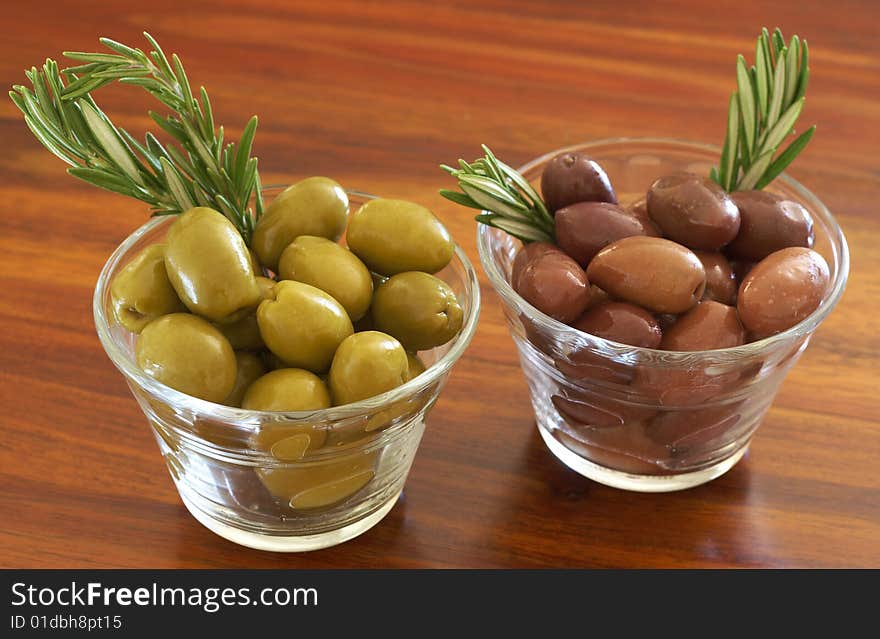 Two jars of green and black olives with stick of rosemary on wooden table background