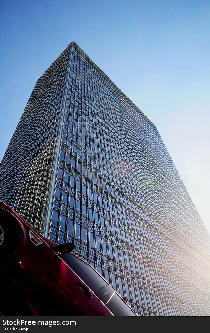 Part of car parked at angle with tall office building in background reaching up to the sky with sun glare on façade. Part of car parked at angle with tall office building in background reaching up to the sky with sun glare on façade.