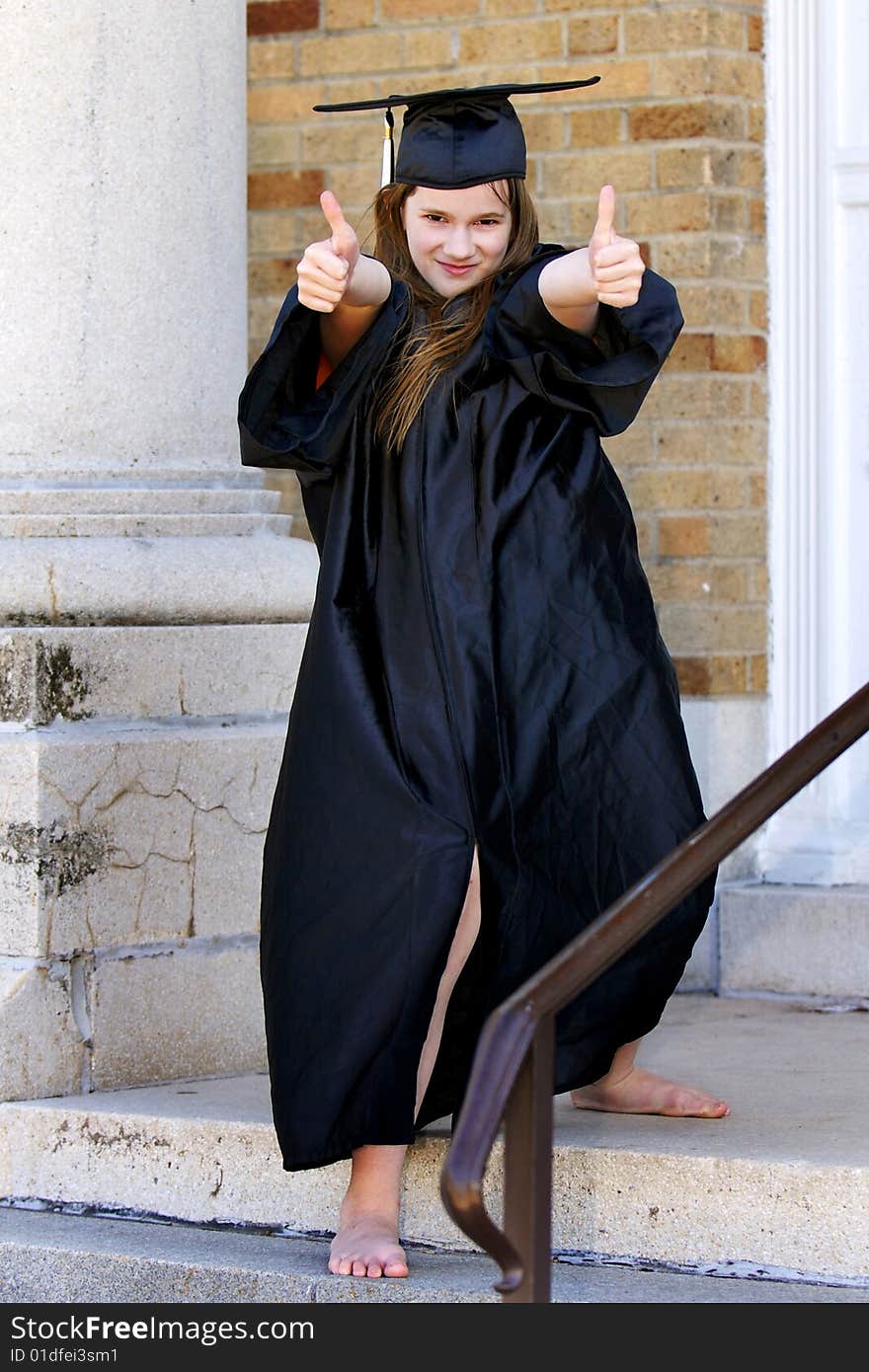 A barefoot middle school graduate in cap and gown, displaying thumbs up on the steps of her school. A barefoot middle school graduate in cap and gown, displaying thumbs up on the steps of her school.