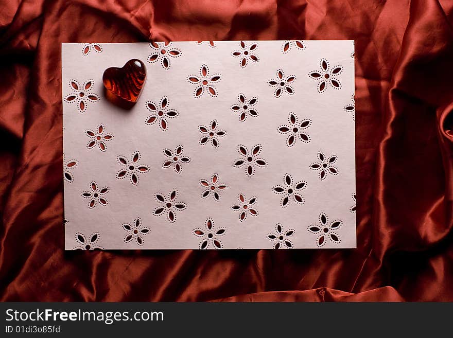 A pink sheet of paper decorated with flowers and a red glass heart, lying on red satin background. A pink sheet of paper decorated with flowers and a red glass heart, lying on red satin background.