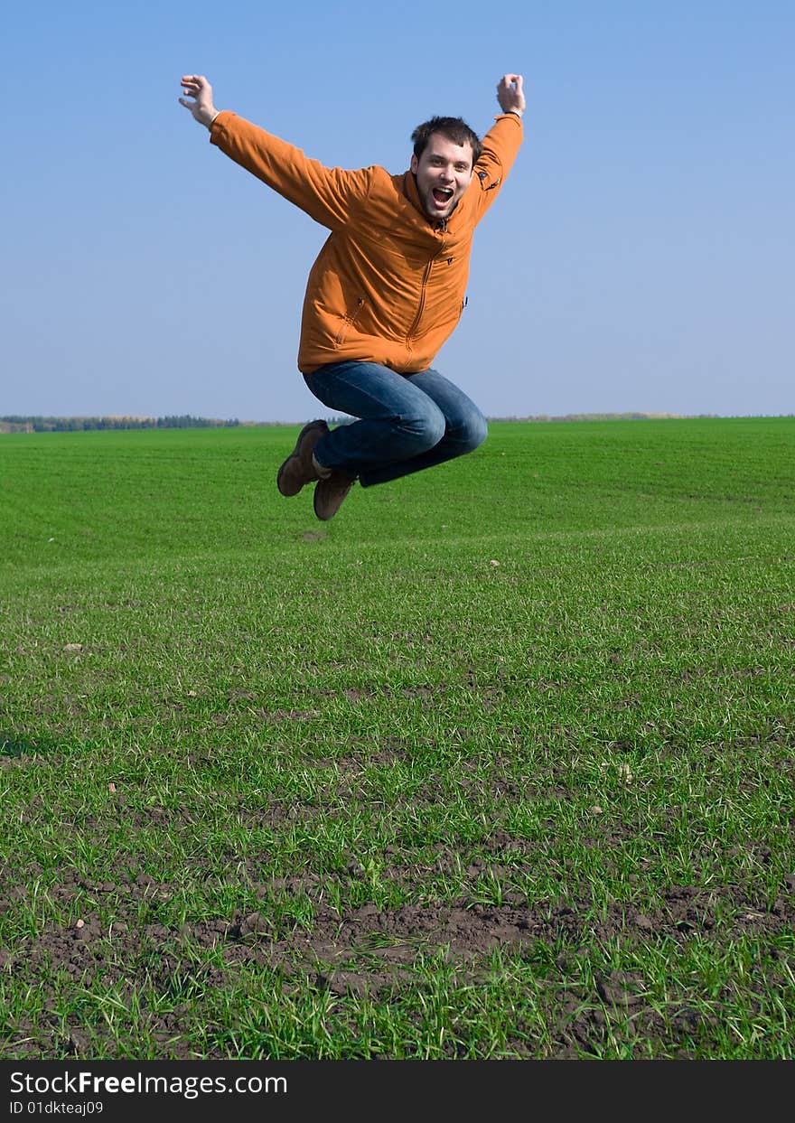 Jumping man in jeans and orange jacket on blue sky and green grass background