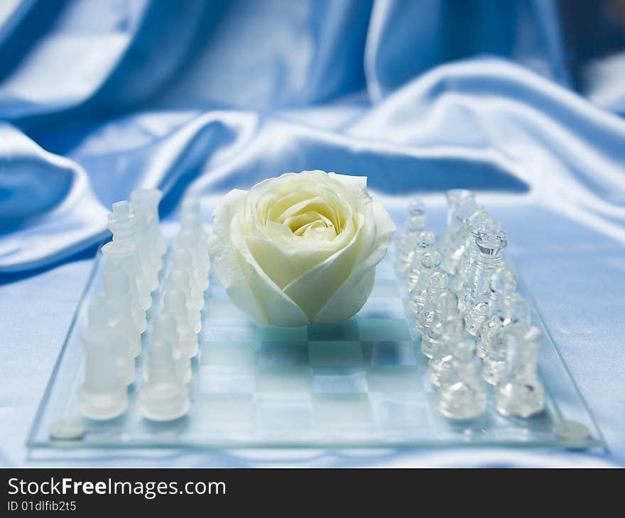 Glass chess board and a white rose in the center symbolizing peace, blue satin in the background. Glass chess board and a white rose in the center symbolizing peace, blue satin in the background.