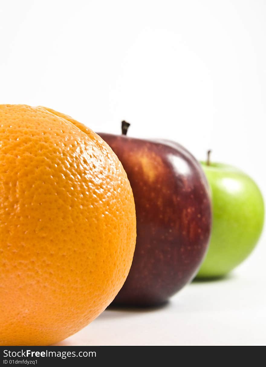 An orange, red apple and green apple lined up showing half of their shapes; the focus is on the orange while the rest are slightly blurred. An orange, red apple and green apple lined up showing half of their shapes; the focus is on the orange while the rest are slightly blurred