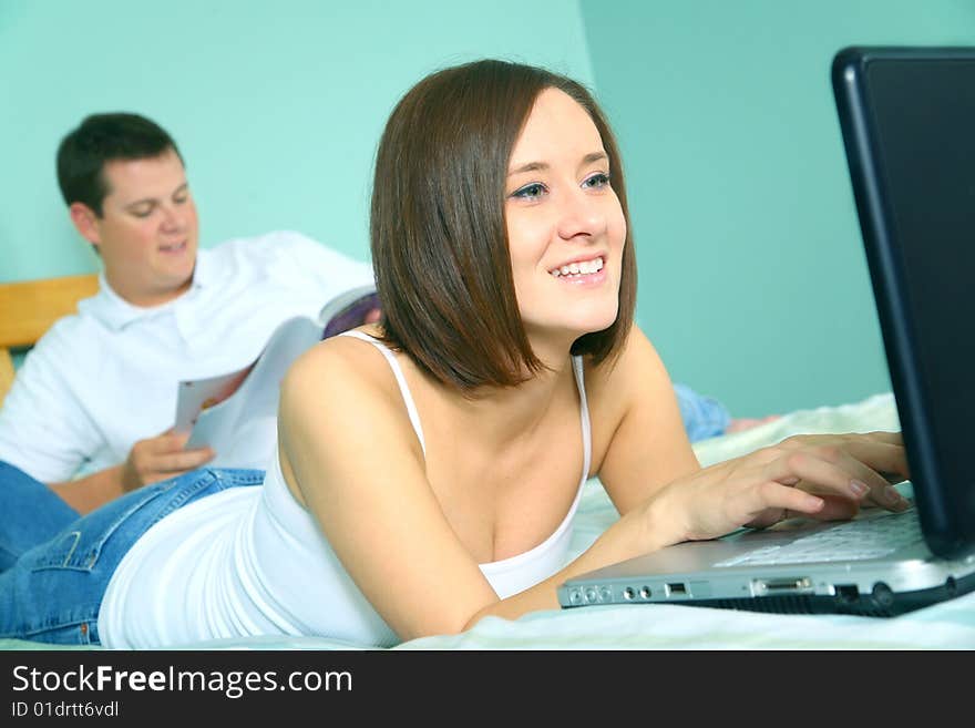 Happy young caucasian female using laptop on bed and a man on the background reading. Happy young caucasian female using laptop on bed and a man on the background reading