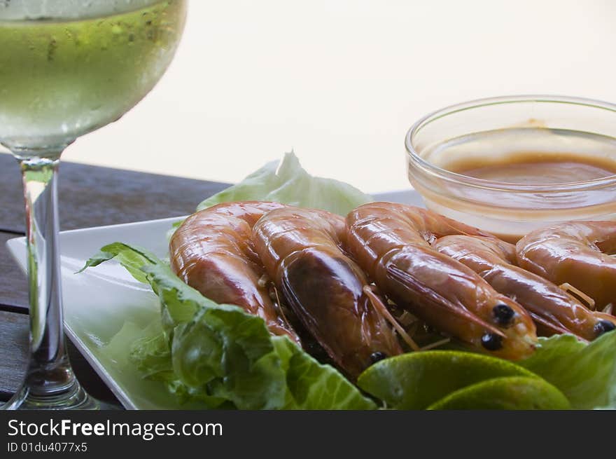 Fresh king prawns placed on a bed of lettuce served with a pink sauce and slices of lemons accompanied by a glass of white wine in an outdoor setting.