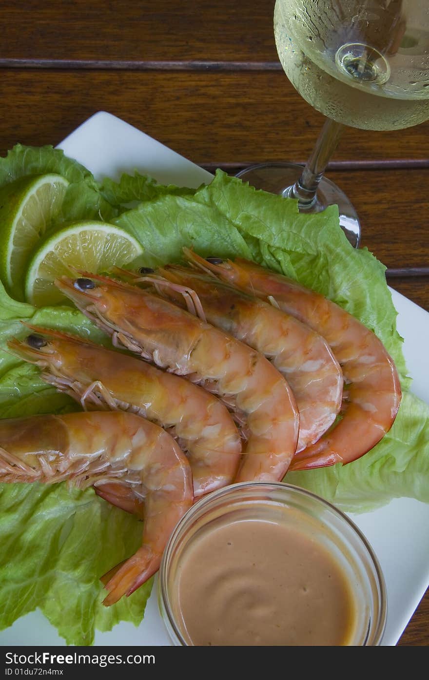 Fresh king prawns placed on a bed of lettuce served with a pink sauce and slices of lemons accompanied by a glass of white wine in an outdoor setting.