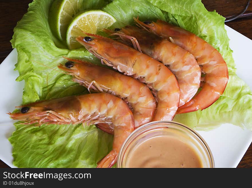 Fresh king prawns placed on a bed of lettuce served with a pink sauce and slices of lemons accompanied by a glass of white wine in an outdoor setting.