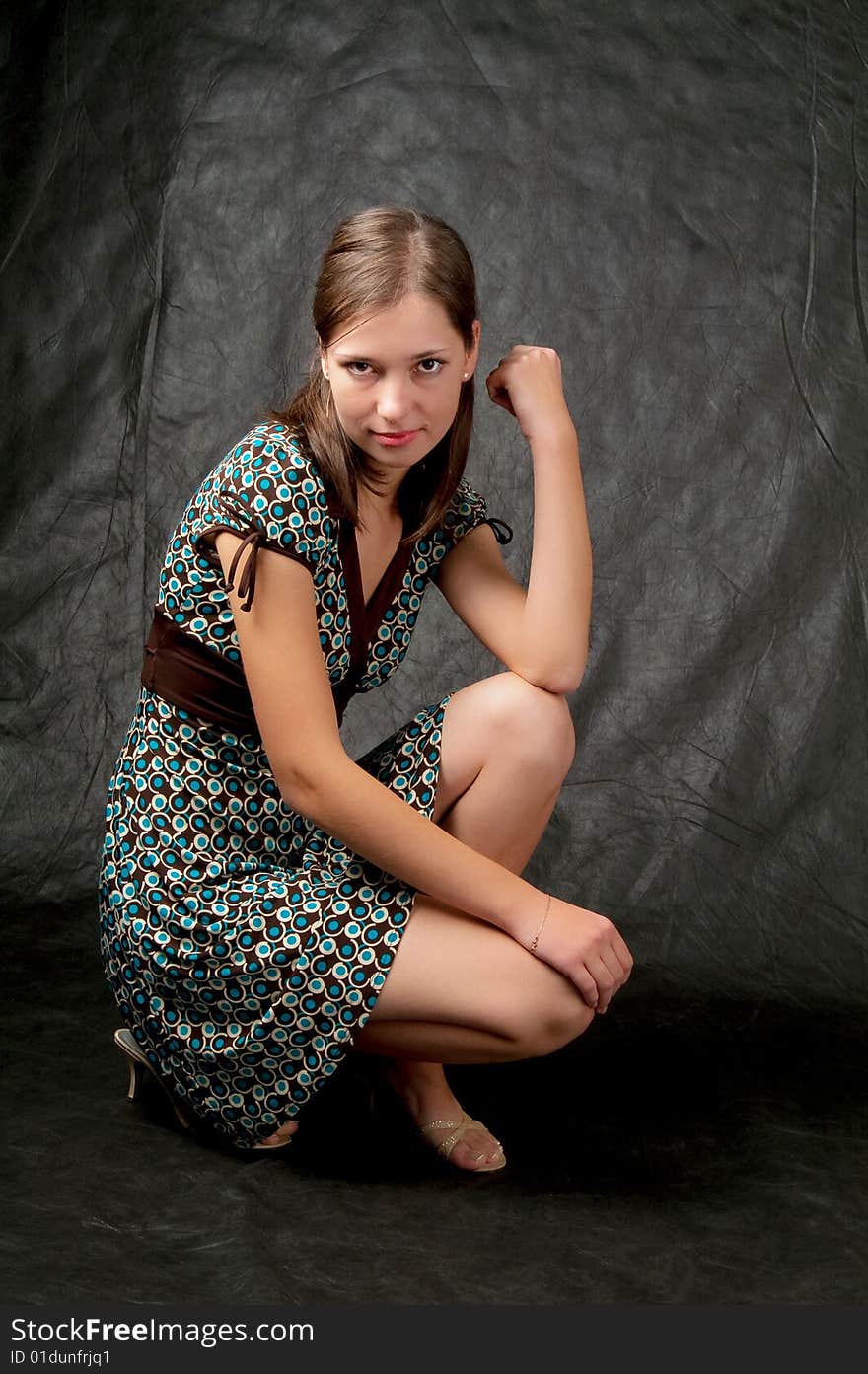 Young girl in color dress with white skin straight dark hair sitting turned left side with head turned with naked legs and free pose isolated on black. Young girl in color dress with white skin straight dark hair sitting turned left side with head turned with naked legs and free pose isolated on black