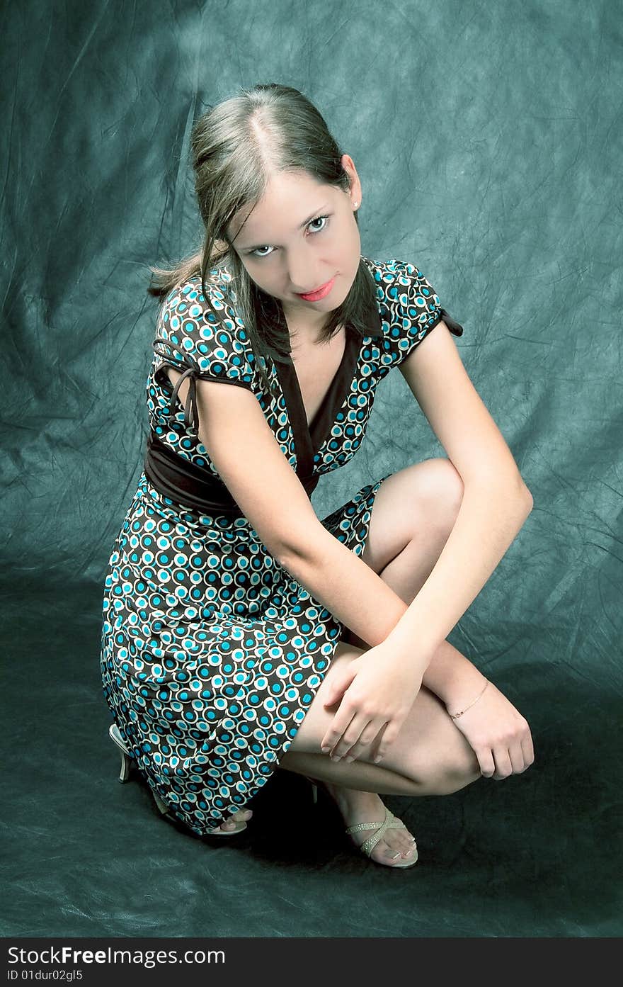 Young white girl in color dress sitting on her hunkers turned slightly smiling with hands crossed in front put on knees isolated on black. Young white girl in color dress sitting on her hunkers turned slightly smiling with hands crossed in front put on knees isolated on black