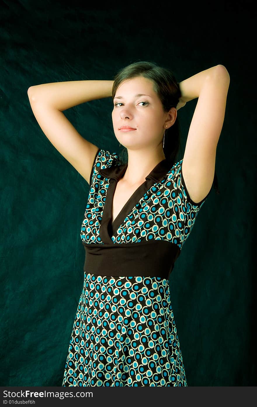 Young caucasian girl with soft skin standing turned with hands up crossed behind head looking left side wearing colored dress with brown belt isolated on black. Young caucasian girl with soft skin standing turned with hands up crossed behind head looking left side wearing colored dress with brown belt isolated on black