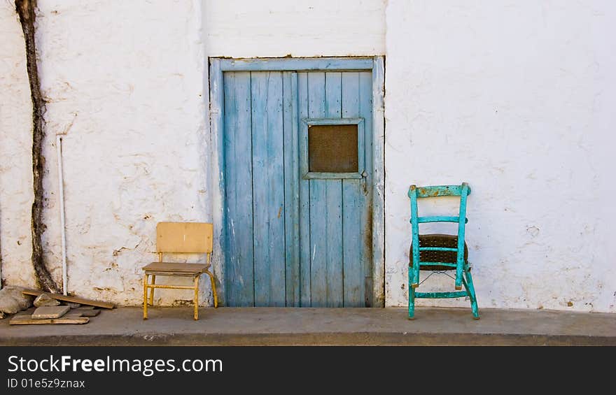 Two old chairs standing on a sidwalk in front of a wall with a blue door. Two old chairs standing on a sidwalk in front of a wall with a blue door