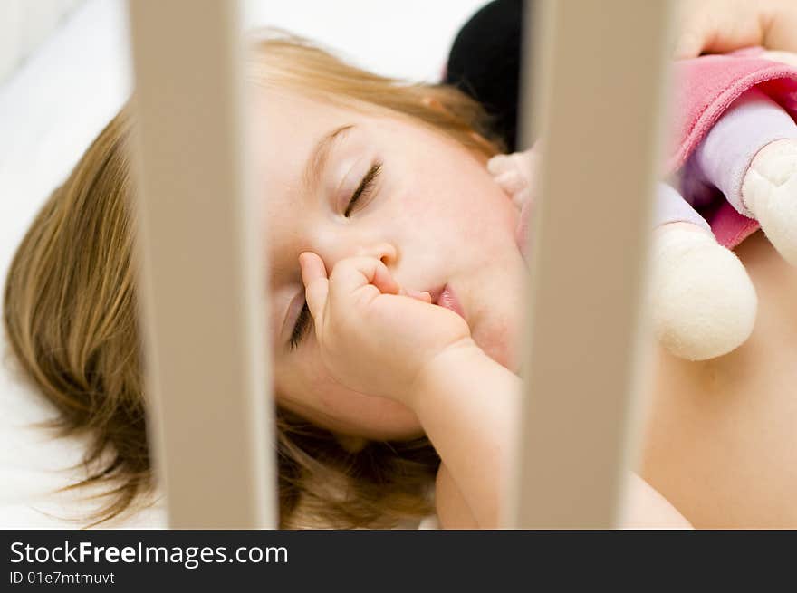 Beautiful baby girl sucking her thumb and hugging her doll in her cot just before bedtime