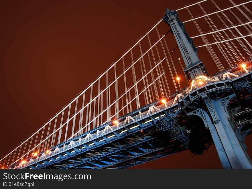 This is a shot of one of the vertical support structures of the Manhattan Bridge. The shot was taken just after sundown. The location was on the Brooklyn side of the Bridge. The location is between the Brooklyn Bridge and looking up from the park area below. The bridge is lit by floodlights provided by the city and appears light blue in the image. The sky has a dark reddish brown tone in the color. Neither the Hudson river or the city of Manhattan are visible in this image. This is a shot of one of the vertical support structures of the Manhattan Bridge. The shot was taken just after sundown. The location was on the Brooklyn side of the Bridge. The location is between the Brooklyn Bridge and looking up from the park area below. The bridge is lit by floodlights provided by the city and appears light blue in the image. The sky has a dark reddish brown tone in the color. Neither the Hudson river or the city of Manhattan are visible in this image.