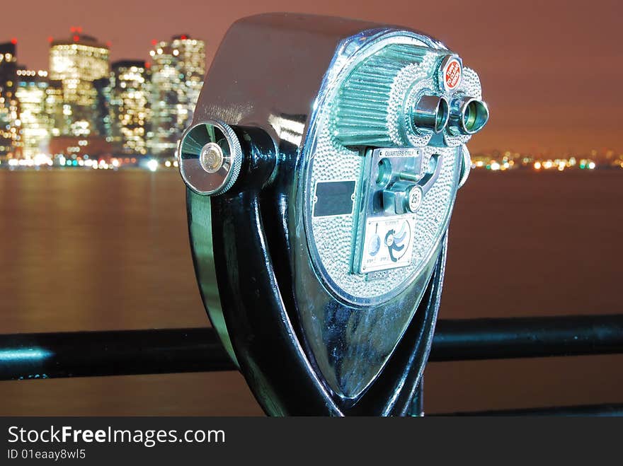 This is a shot of a coin operated viewfinder. This viewfinder is found on the Jersey side of the Hudson river. In the background the city of Manhattan can be seen. The subject is not the city but the finder itself. The image was captured at night. Lighting was provided by some local street lights. The finder is ever slightly tilted back. It is rotated on its axis so the back and part of one side is seen. A very small section on top of the finder is seen. The color of the finder is silver. The background has a brownish look and shows the city and part of the river in the image. A protective fall off rail can be seen running the width of the image in the lower third. This is a shot of a coin operated viewfinder. This viewfinder is found on the Jersey side of the Hudson river. In the background the city of Manhattan can be seen. The subject is not the city but the finder itself. The image was captured at night. Lighting was provided by some local street lights. The finder is ever slightly tilted back. It is rotated on its axis so the back and part of one side is seen. A very small section on top of the finder is seen. The color of the finder is silver. The background has a brownish look and shows the city and part of the river in the image. A protective fall off rail can be seen running the width of the image in the lower third.