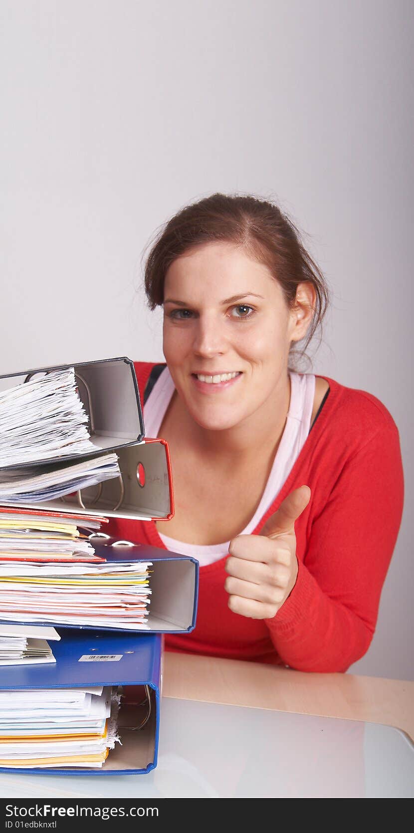 A young and beautiful female worker behind a lot of paperwork. She gives a thumb up sign. A lot of copyspace.