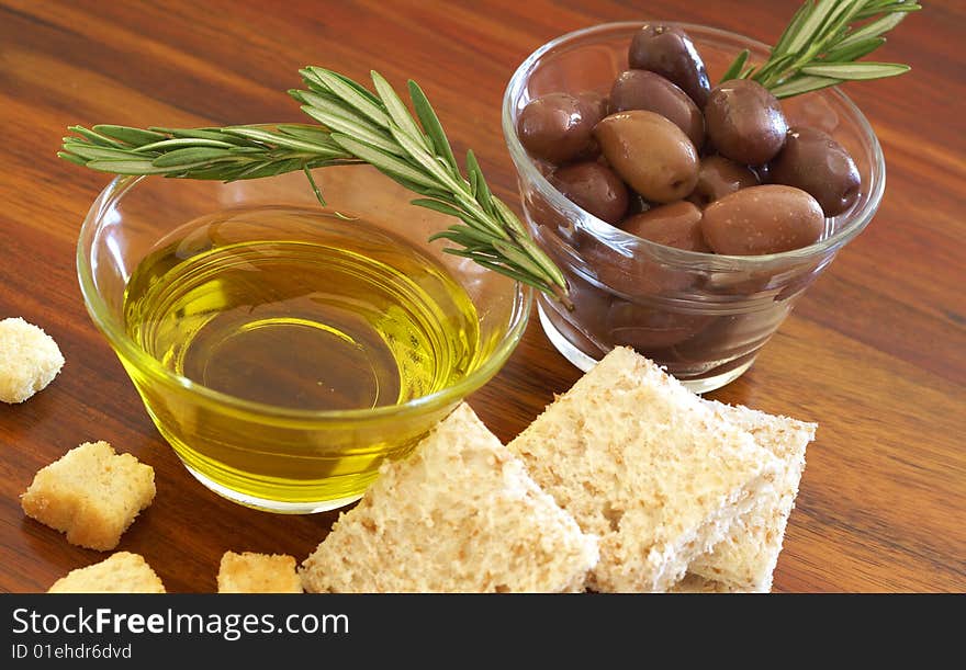 Two jars of black olives with stick of rosemary, olive oil, slices of bread and croutons on wooden table background. Two jars of black olives with stick of rosemary, olive oil, slices of bread and croutons on wooden table background